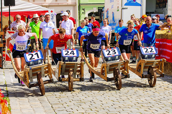 sportlich angezogene Menschen mit Startnummern, Frauen und Männer, schieben etwas, was aussieht wie eine Schubkarre aus Holz. Hinter ihnen stehen Zuschauer, die dem ganzen gespannt folgen.