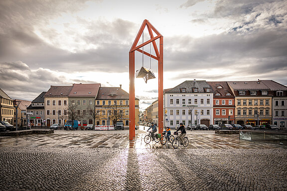 Zu sehen ist ein Marktplatz. Im Hintergrund reihen sich Stadthöuser in verschiedenen Farben auf. Im Vordergrund sieht man drei Fahrradfahrer. Es hat gerade geregnet und die Sonne bricht durch die Wolkendecke. Im Zentrum steht der Mediaturm aus roten Stahl. Er gleicht mehreren Dreiecken, die sich kreuzen. An diesen Dreieecken hängt an Stahlseilen ein Gebilde, welches einem Stern ähnelt.