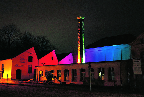Leuchtender Schornstein links im Bils die beleuchtete Schettdach-Halle, mittig der beleuchtete Schornstein, im Hintergrund das Aulagebäude der Oberlausitzer Webschule