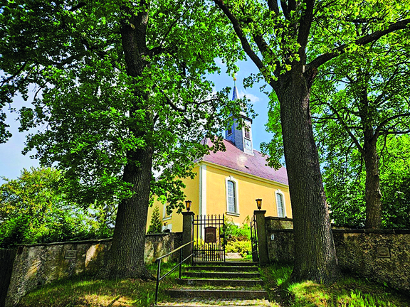 Die Kirche ist eine Station auf der Lauschtour Zu sehen ist eine Barockkirche mit einer gelben Fassade, langgestreckten Barockfenstern und einem roten Walmdach. Außerdem schaut ein Turm aus dem Dach hervor. Im Vordergrund sieht man ein schmiedeisernes Tor, welches zur Kirche führt. Das Tor wird flankiert von zwei großen Bäumen, an denen grüne Blätter hängen.