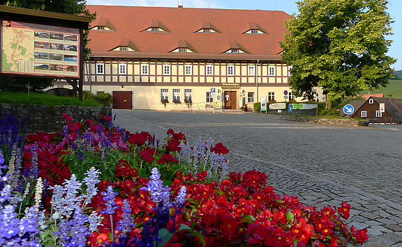 Naturparkhaus Zittauer Gebirge rote und lilafarbene Blumen im Vordergrund des Bildes, linkst steht eine Ortstafel mit Informationen, den gesamten oberen Bildteil dominiert das Naturparkhaus Zittauer Gebirge, ehemaliger Niederkretscham und größtes Umgebindehaus
