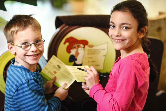 Kinder mit Rangerpass ein etwa achtjähriger Junge und ein neunjähriges Mädchen halten den Fragenzettel für den Junior Ranger Pass in den Händen, stehen vor einem Querx und seiner Frage und blicken lächelnd in die Kamera